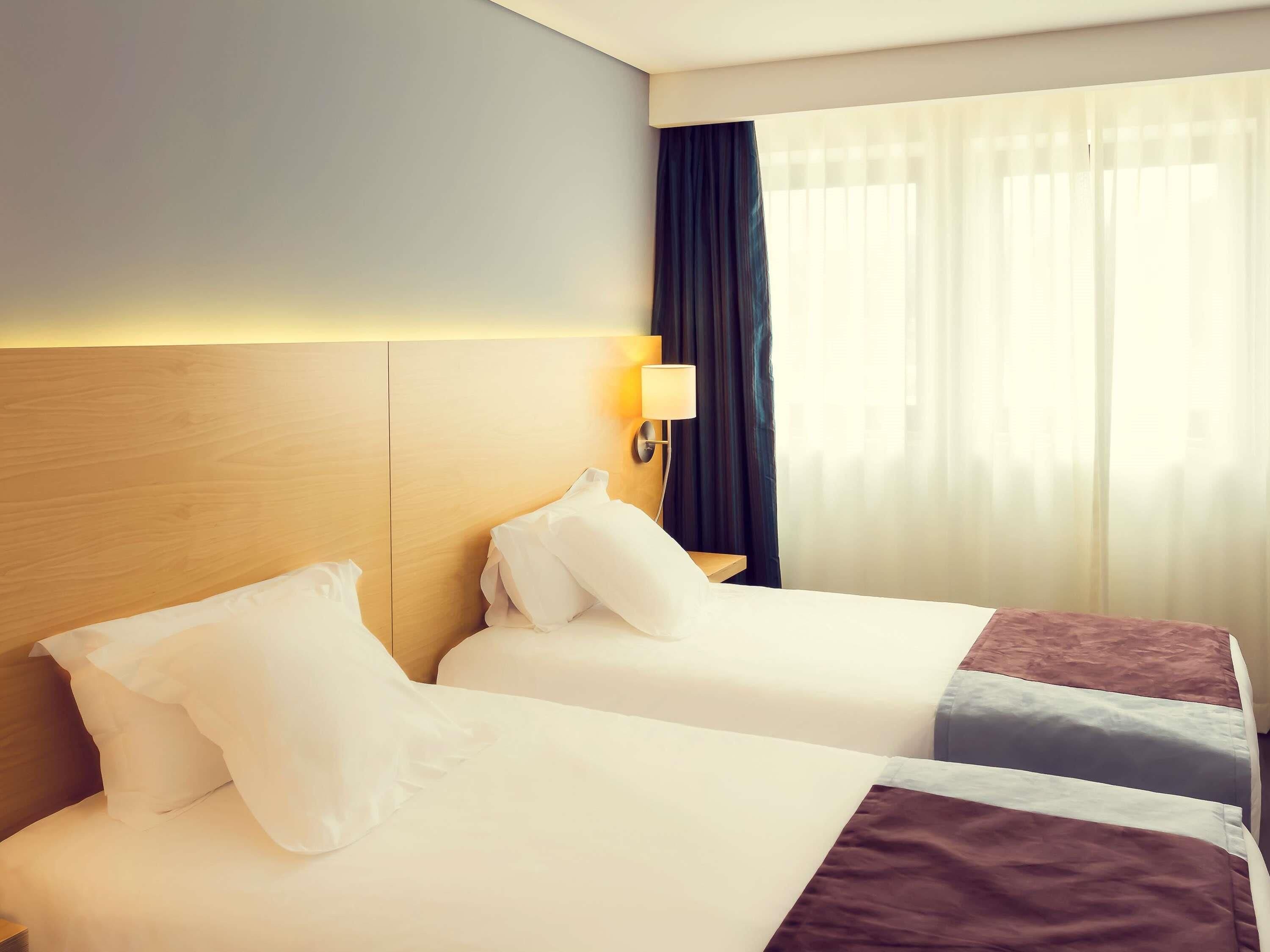 Une chambre d’hôtel avec deux lits, une tête de lit en bois, une lampe de chevet et une fenêtre avec des rideaux.