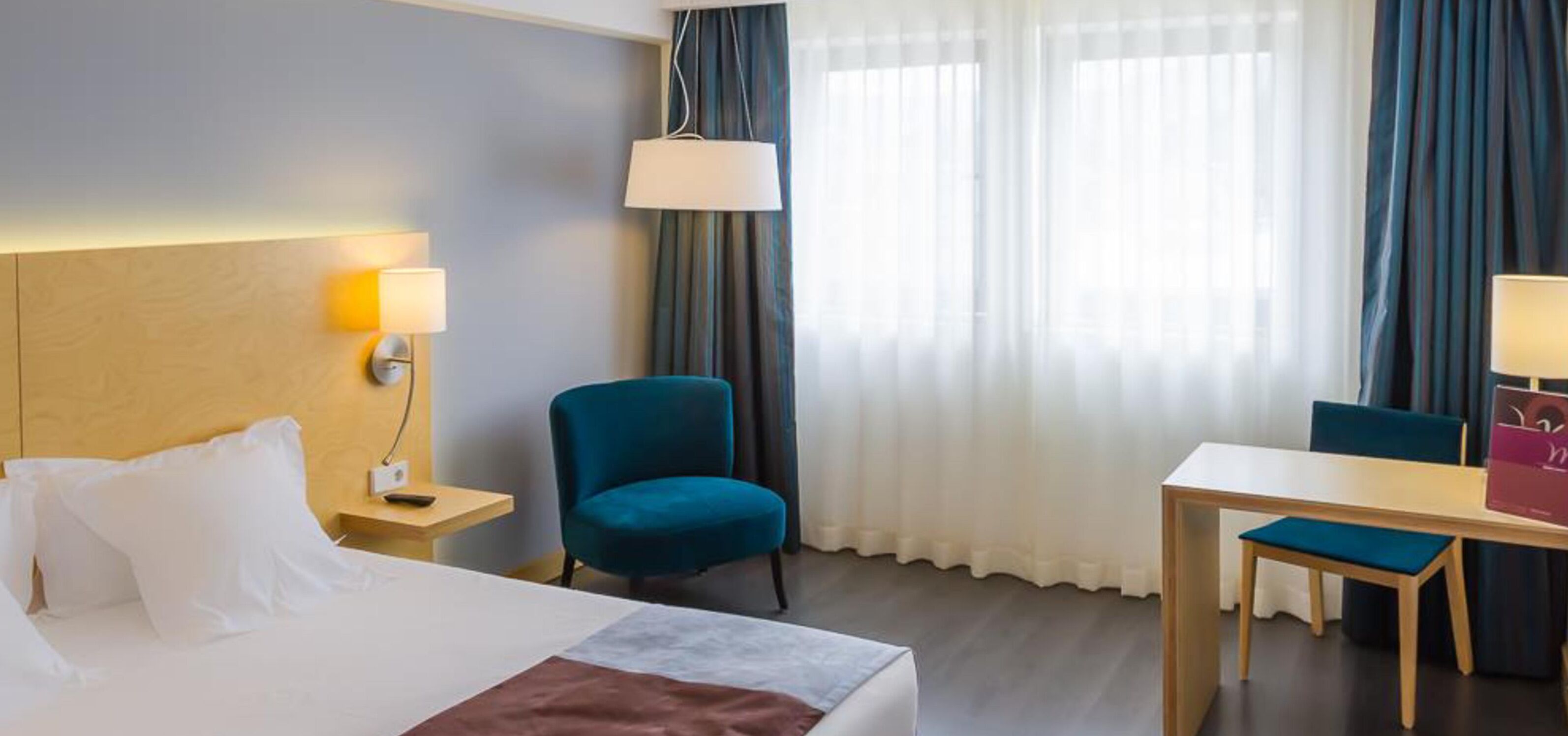 A hotel room with a bed, a desk, a chair, a lamp, and a window with curtains.