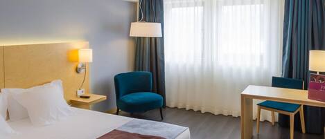 A hotel room with a bed, a desk, a chair, a lamp, and a window with curtains.