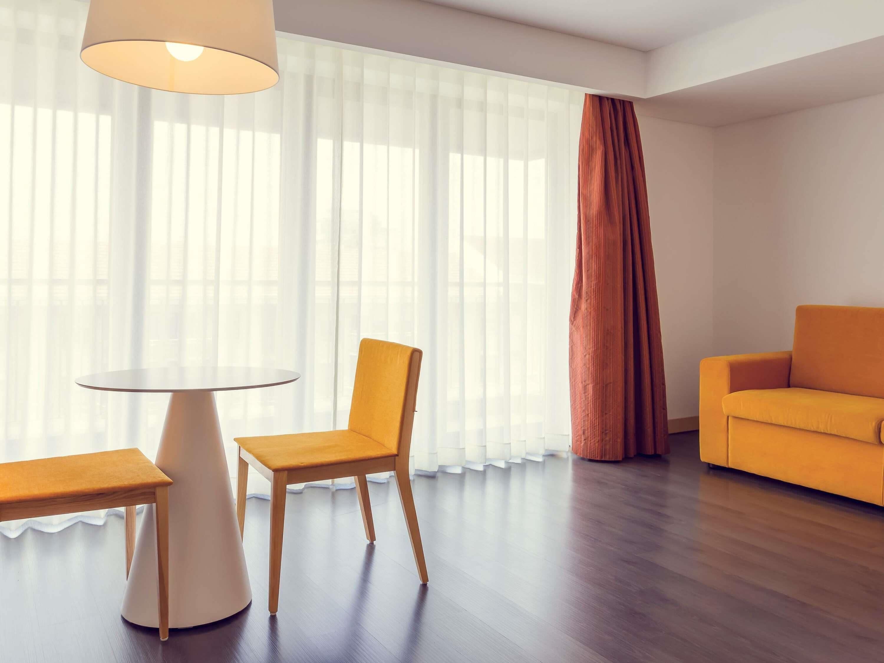 A modern living room with a round table, a wooden chair, a sofa, and a large window with sheer curtains.