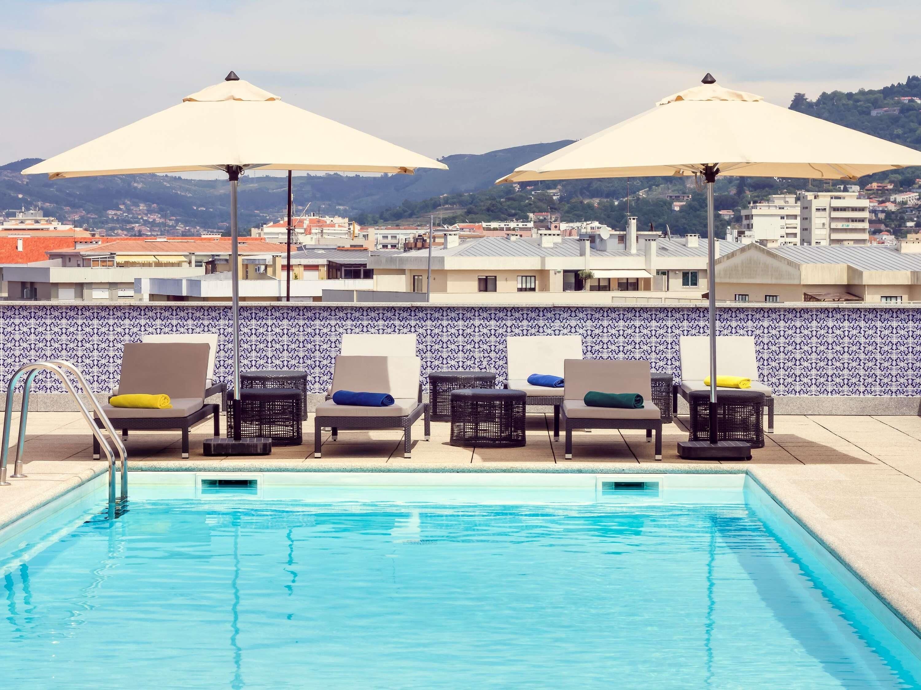 Una piscina en la azotea con sillones de descanso y sombrillas, una pared de azulejos y vistas a la ciudad.