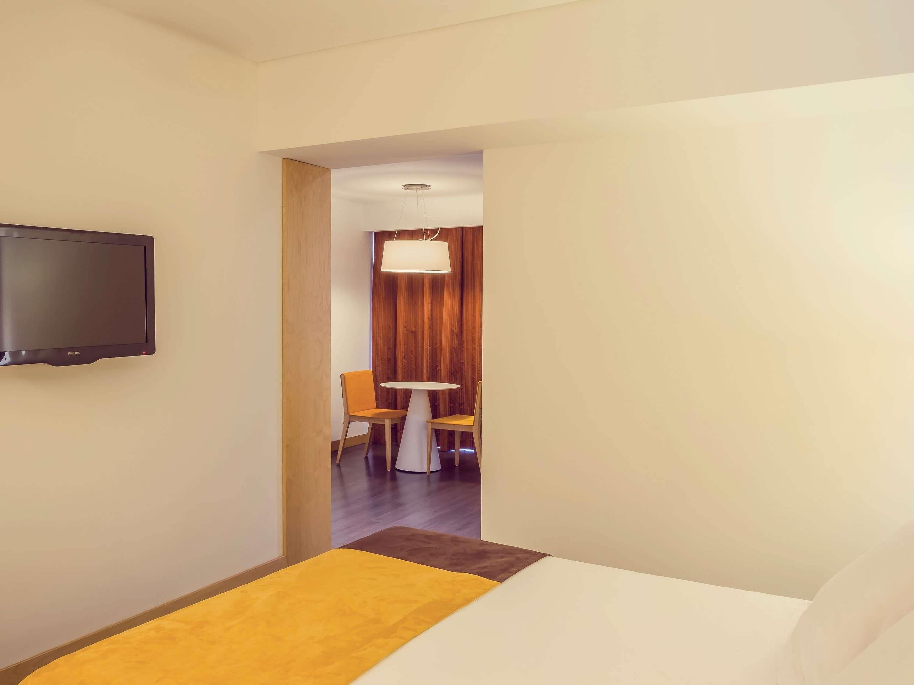 Quarto de hotel com uma cama, televisão, uma mesinha e uma cadeira.
