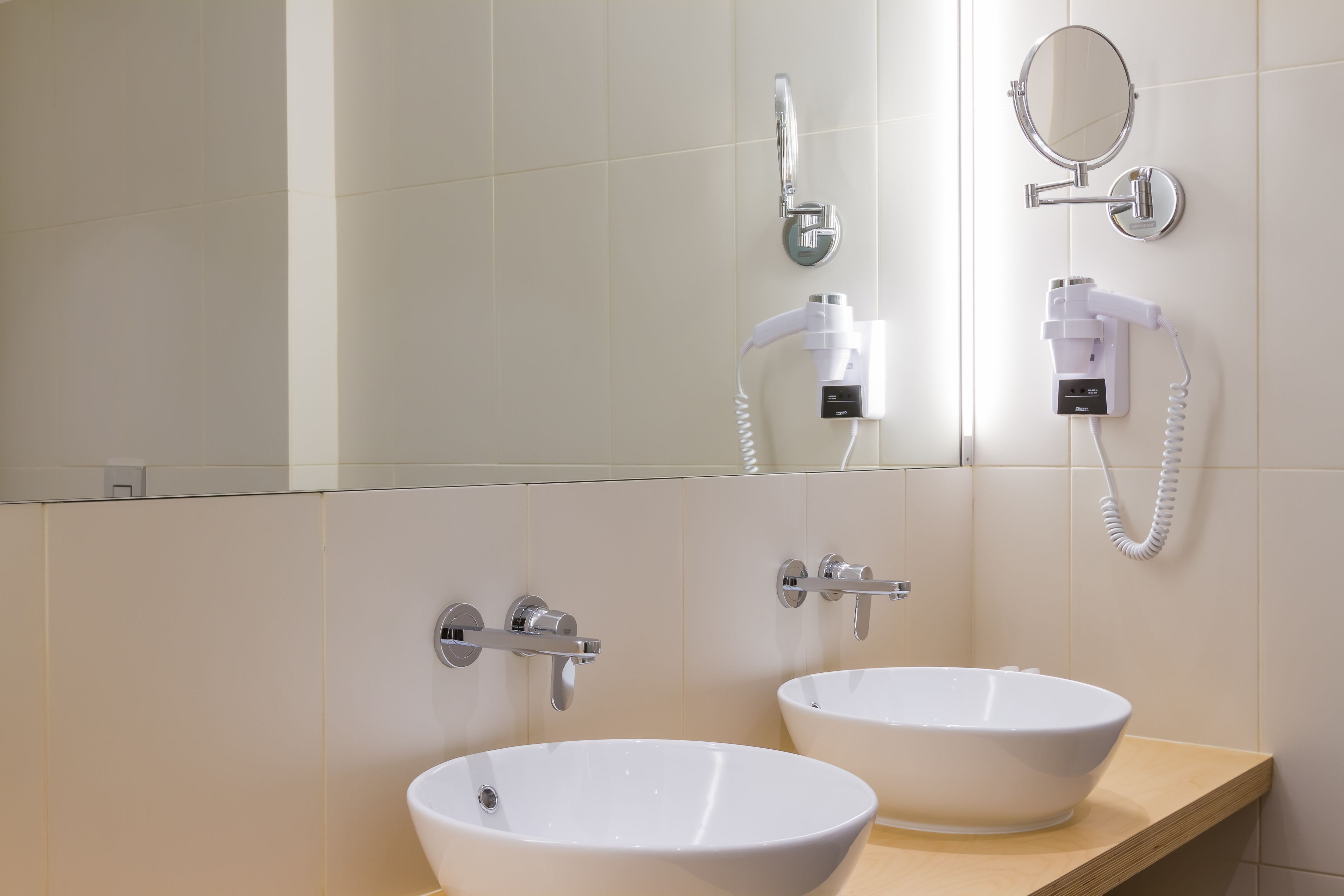 Un baño moderno con dos lavamanos de cerámica blanca, un cabezal de ducha de mano, un espejo montado en la pared y un secador de pelo.