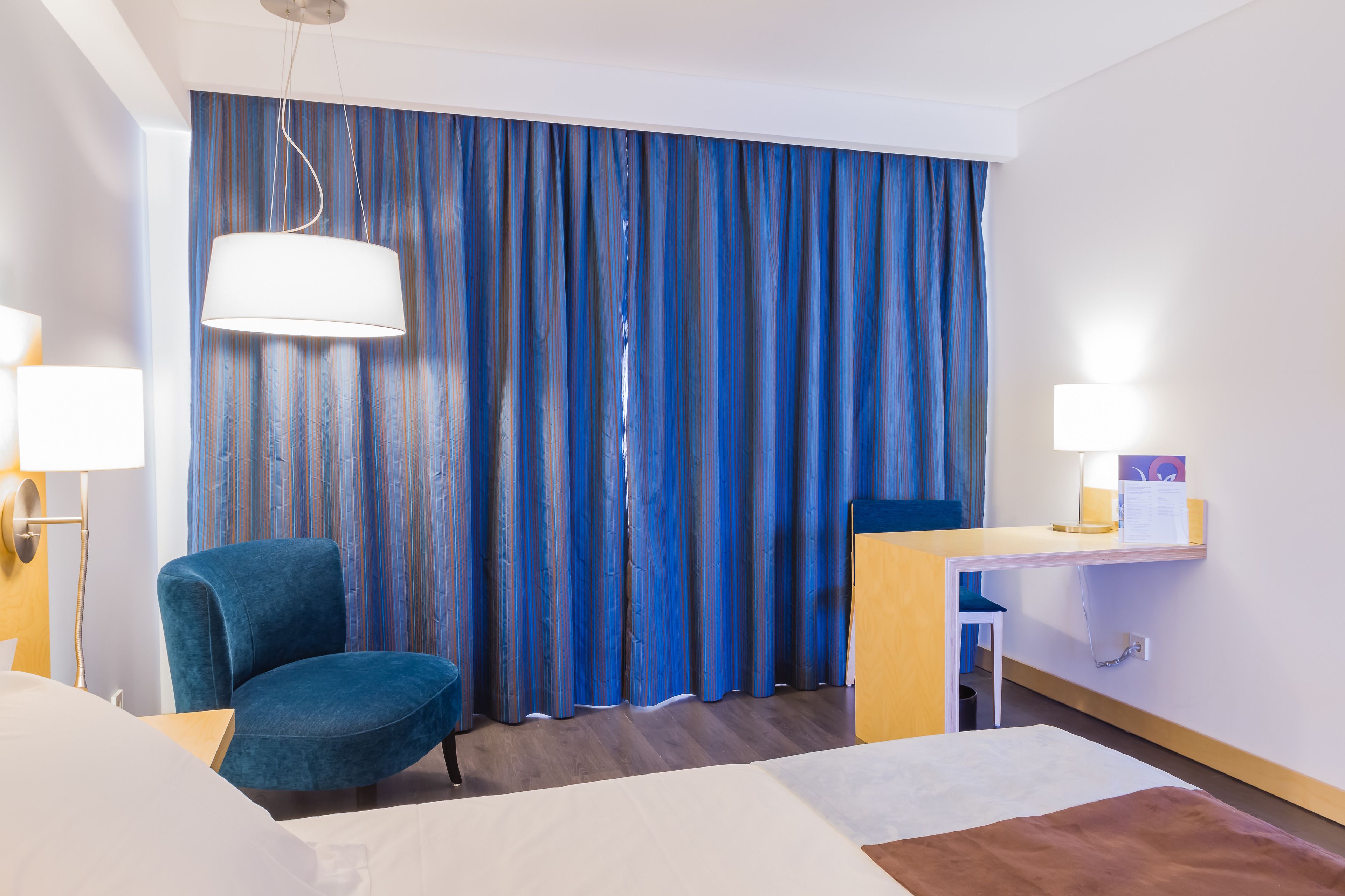 Quarto de hotel com uma cama, uma escrivaninha, uma cadeira e uma cortina.