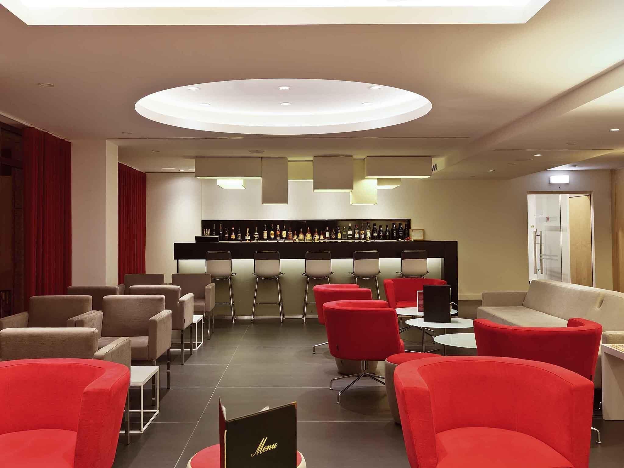 Zona bar dal design moderno, con una lampada a soffitto circolare, un bancone bar con diversi sgabelli e una disposizione di sedute nei toni del rosso e del beige.