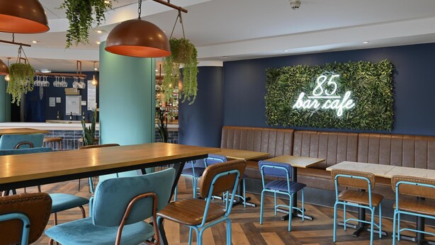 Interior moderno de bar cafetería con mesas de madera y sillas azules, un letrero de neón que dice '85 bar cafe', y plantas colgantes.