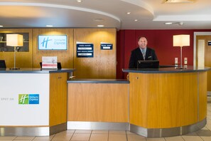 A Holiday Inn Express reception desk with a wooden finish, a digital sign displaying the hotel name, and a "Room Tariff" sign. - Holiday Inn Express Swansea - East by IHG (Neath)