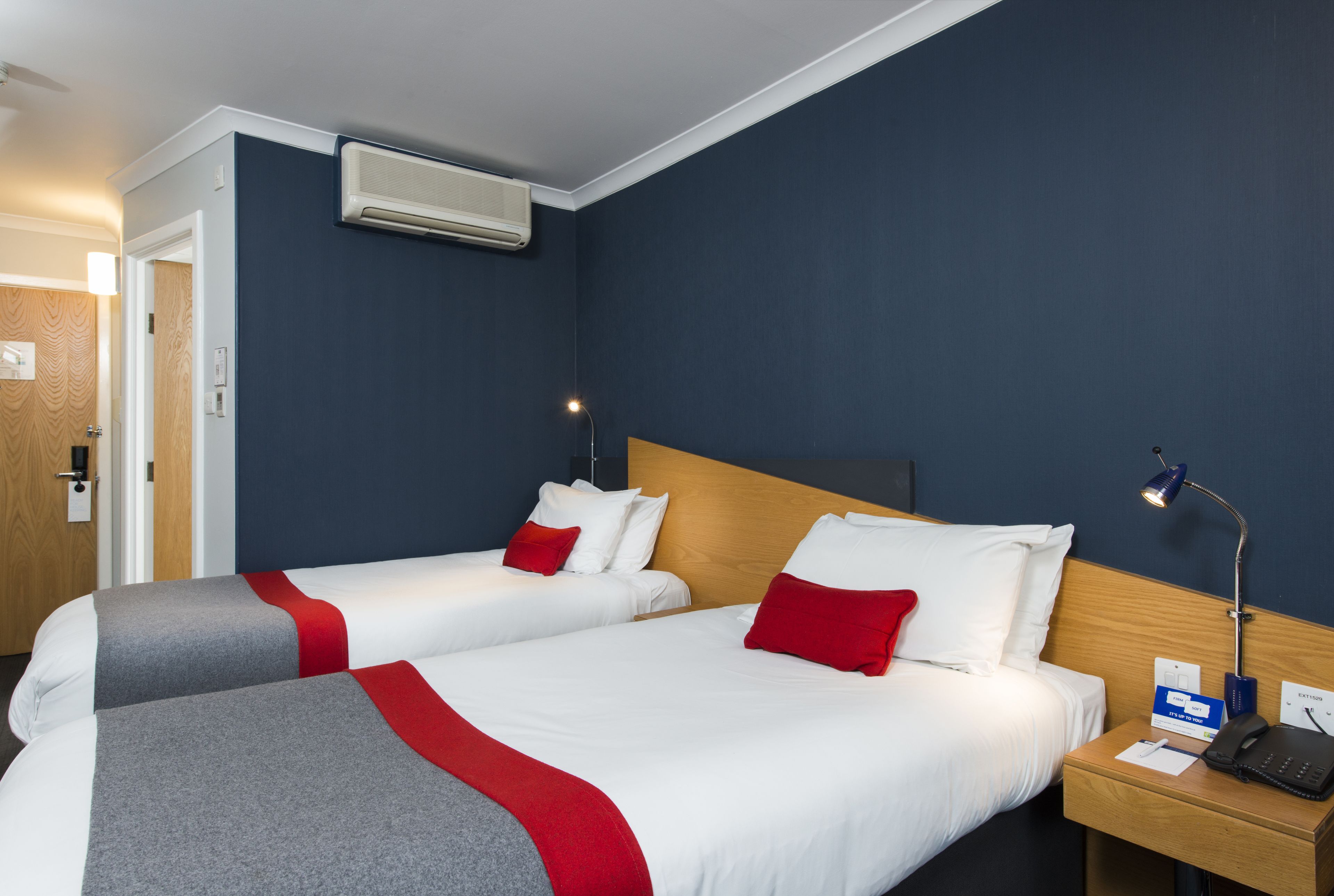 A hotel room with two beds, each with white bedding and red accents, a wooden headboard, a bedside table with a lamp, and an air conditioning unit on the wall.