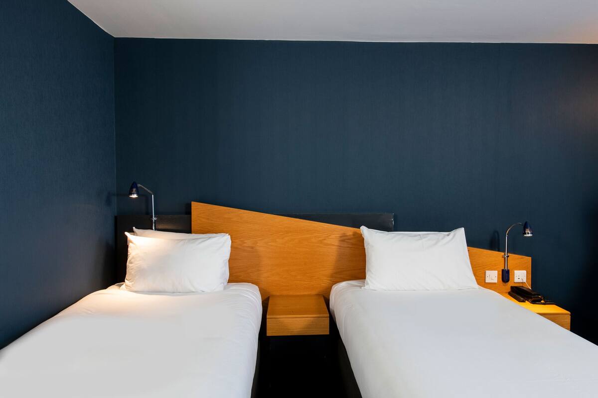 A hotel room with two single beds, wooden headboards, white bedding, and bedside lamps.