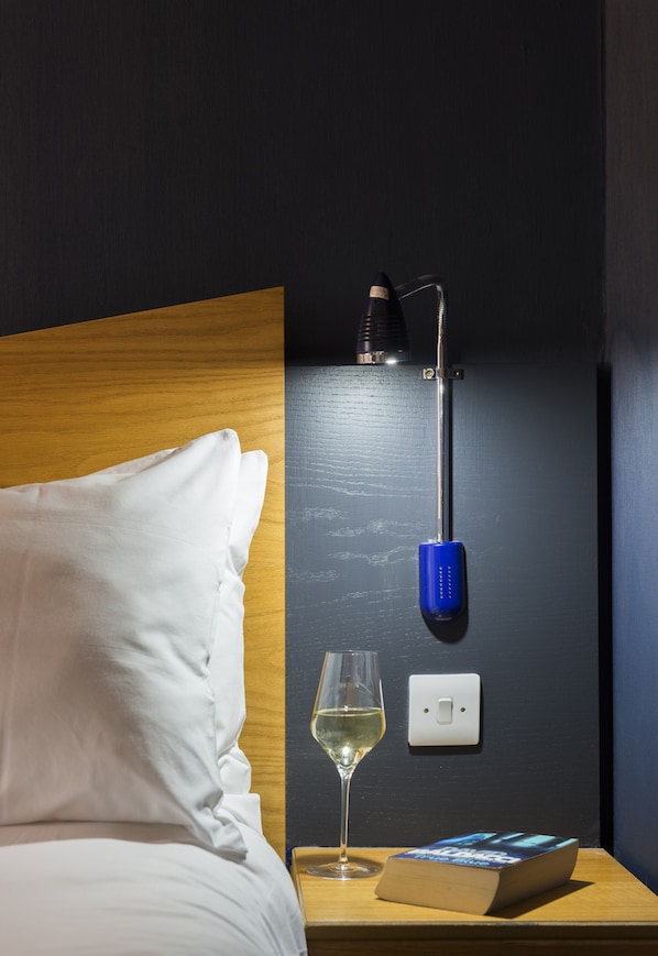 A neatly made bed with white linens, a bedside table with a wine glass and a book, and a wall-mounted lamp with a blue base.