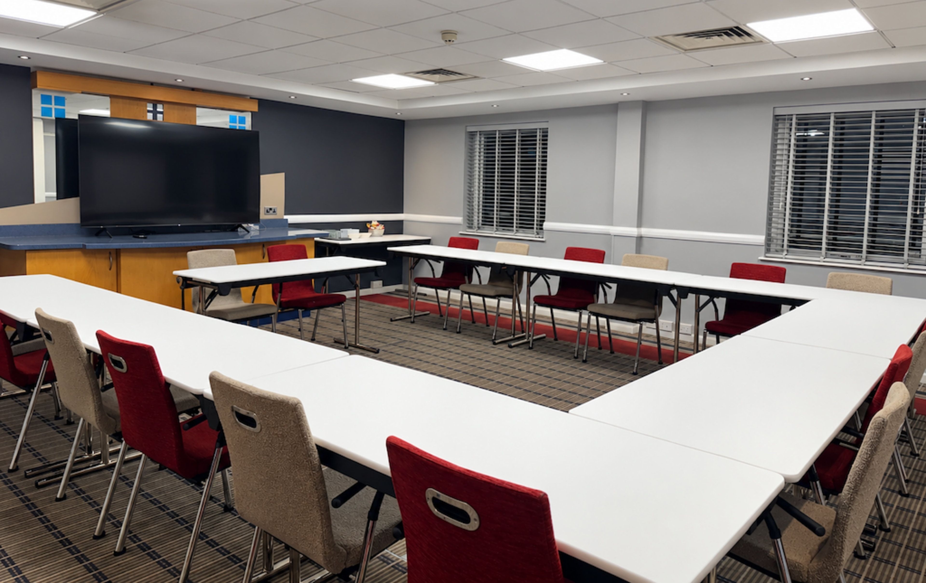 Un salón de conferencias con una larga mesa, sillas rojas, una pantalla grande y ventanas con persianas.