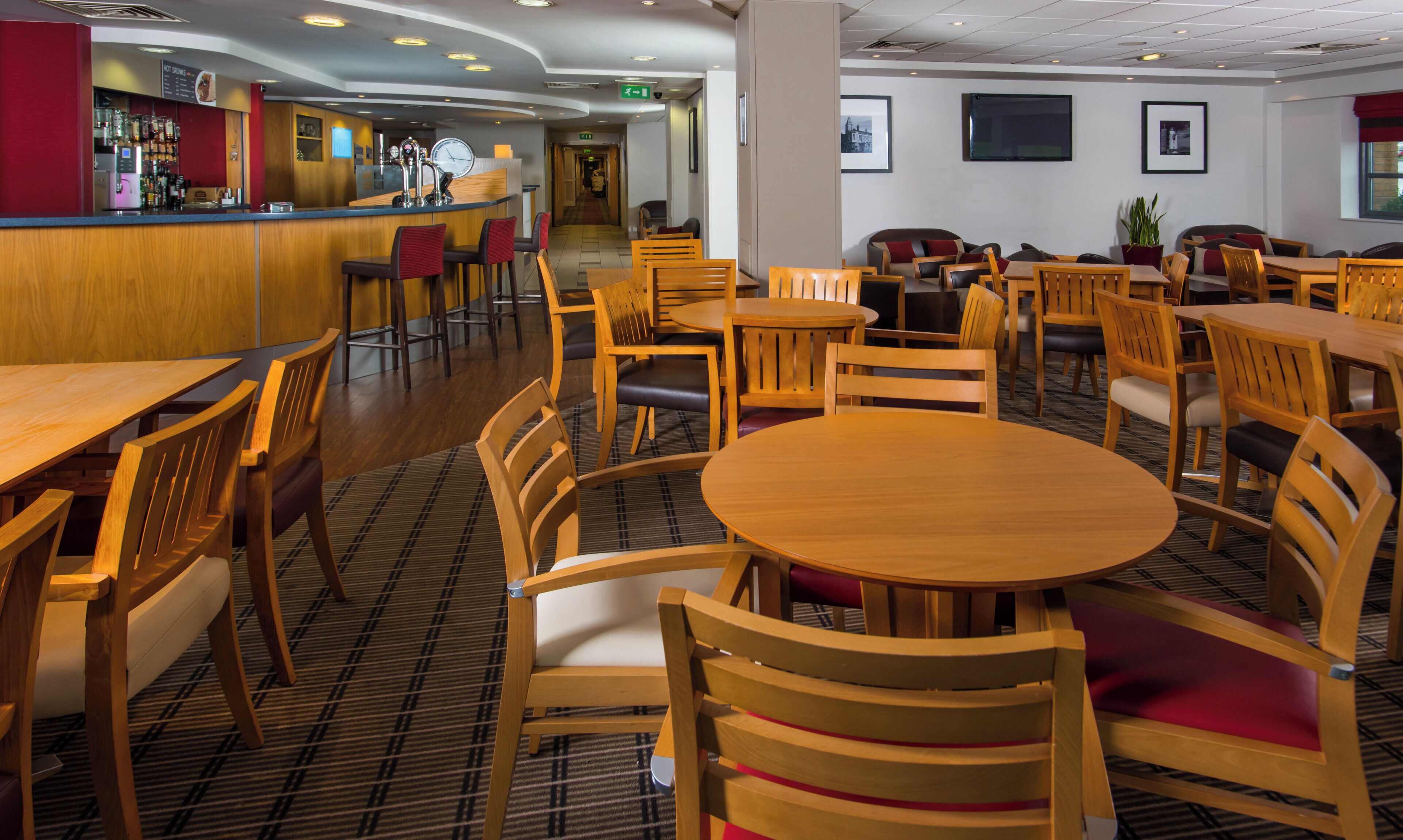 A hotel dining area with wooden tables and chairs, a bar area with a counter and stools, and framed pictures on the walls.