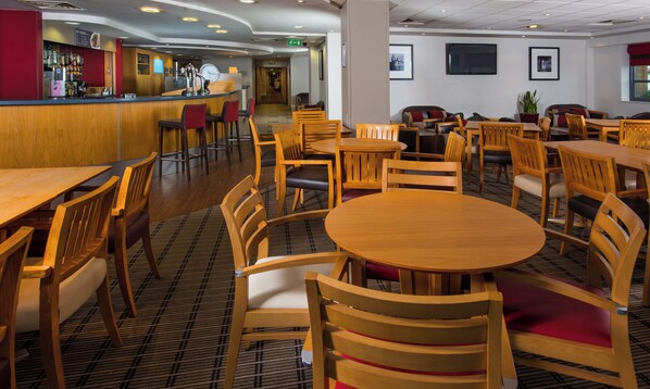 A hotel dining area with wooden tables and chairs, a bar area with a counter and stools, and framed pictures on the walls.