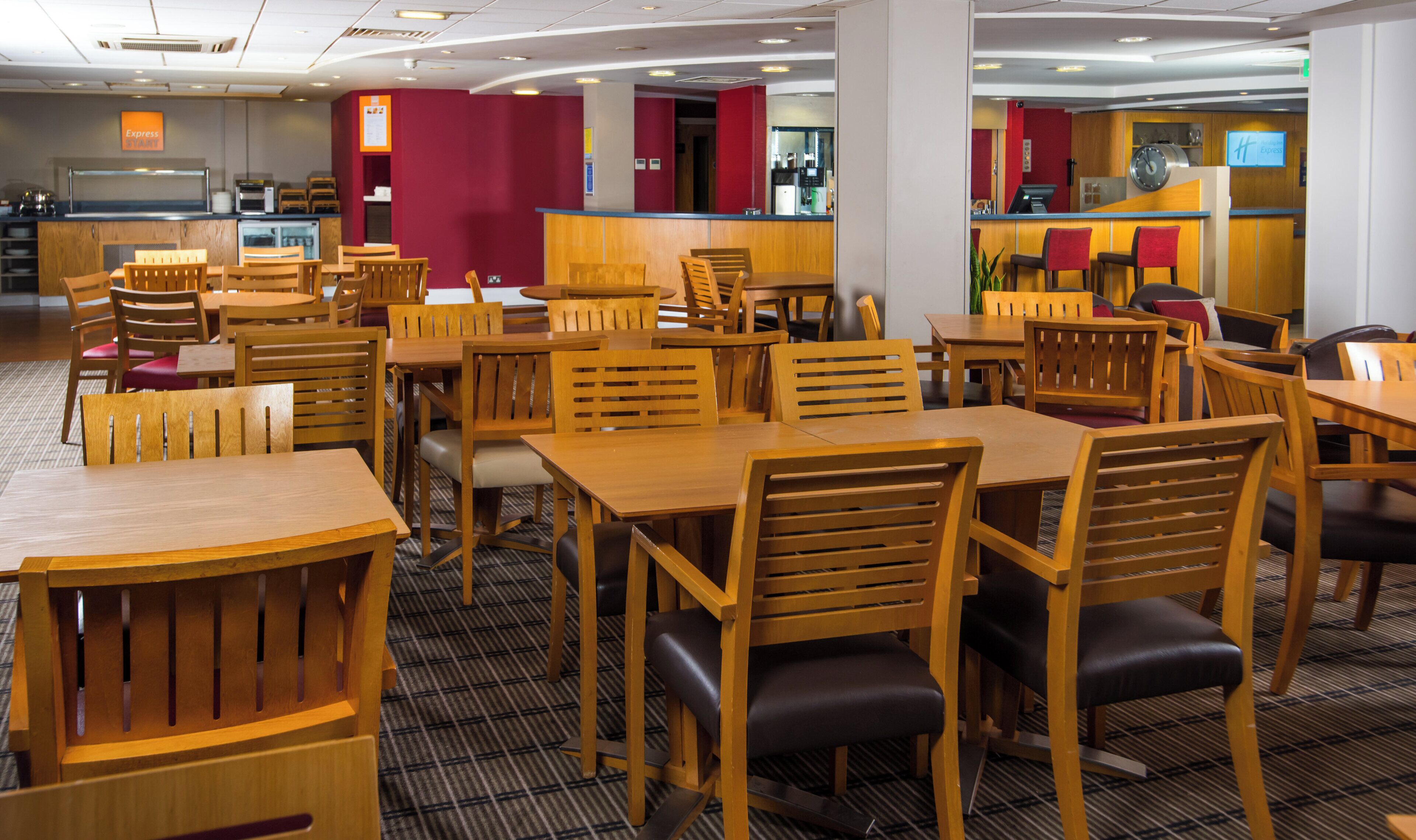 A hotel restaurant with wooden tables and chairs, a red accent wall, and a self-service area in the background.