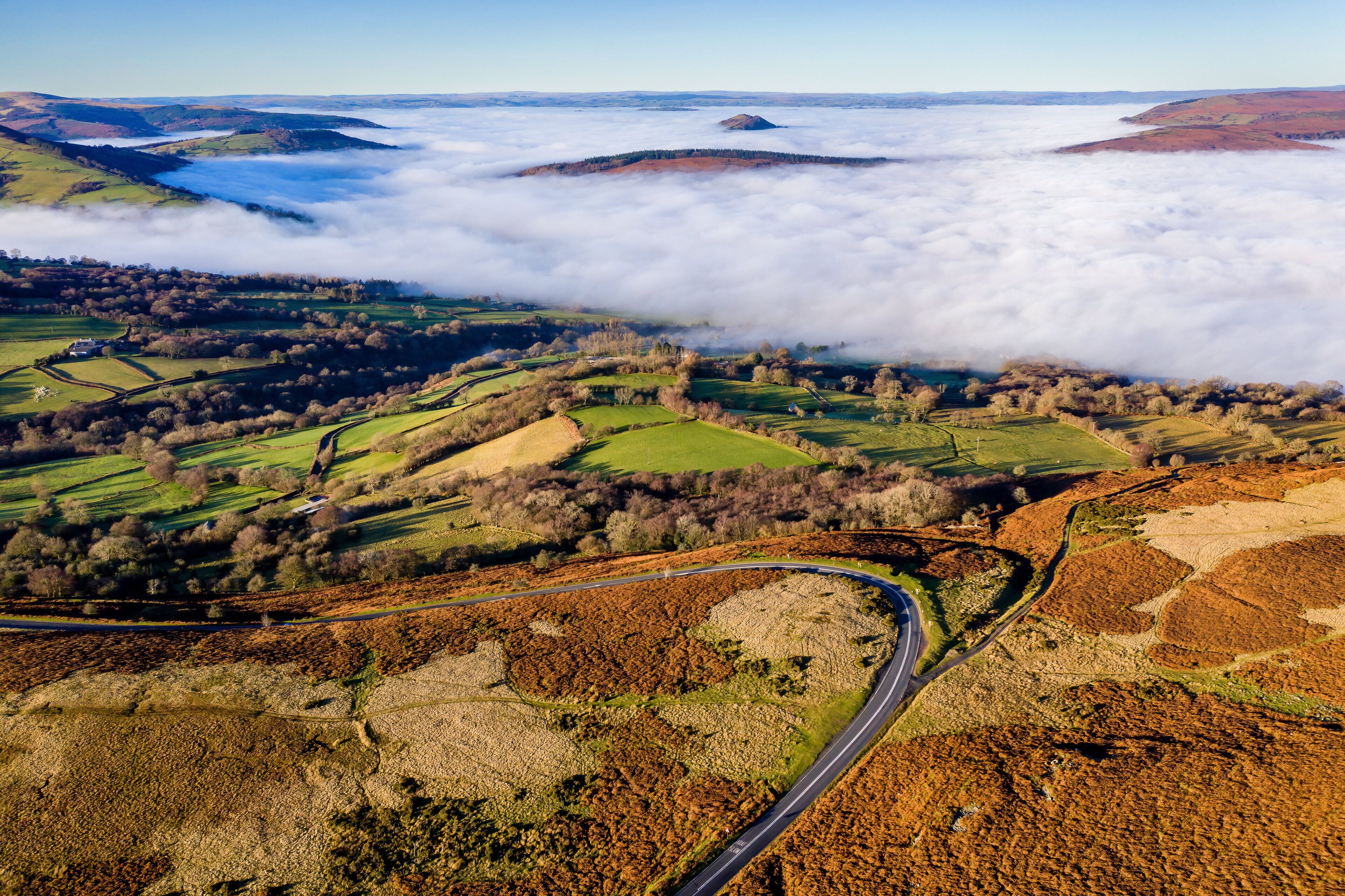 Eine kurvenreiche Straße durch eine hügelige Landschaft mit Feldern und einem Gewässer in der Ferne.