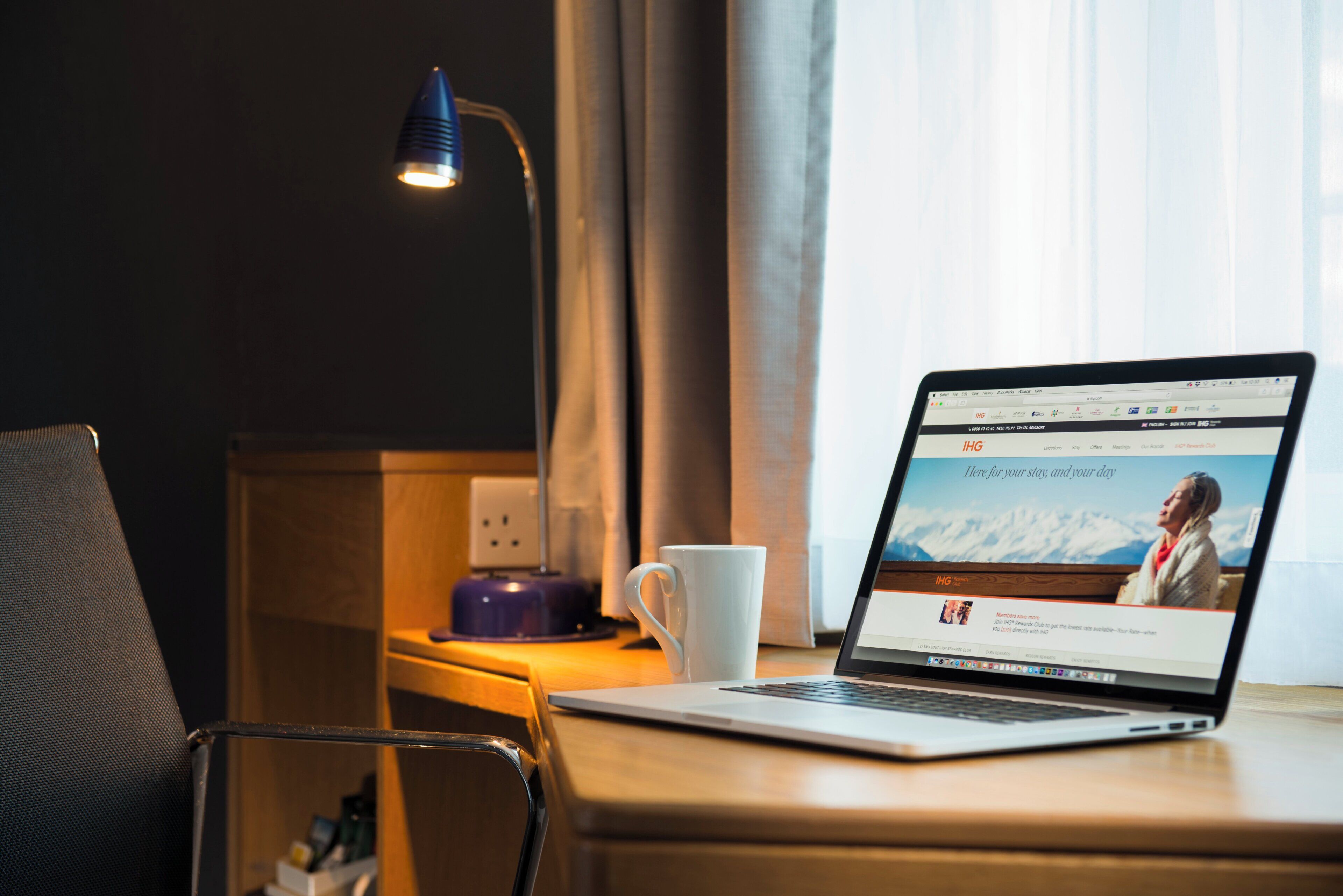 A laptop on a wooden desk with a white mug, a lamp, and a power outlet.