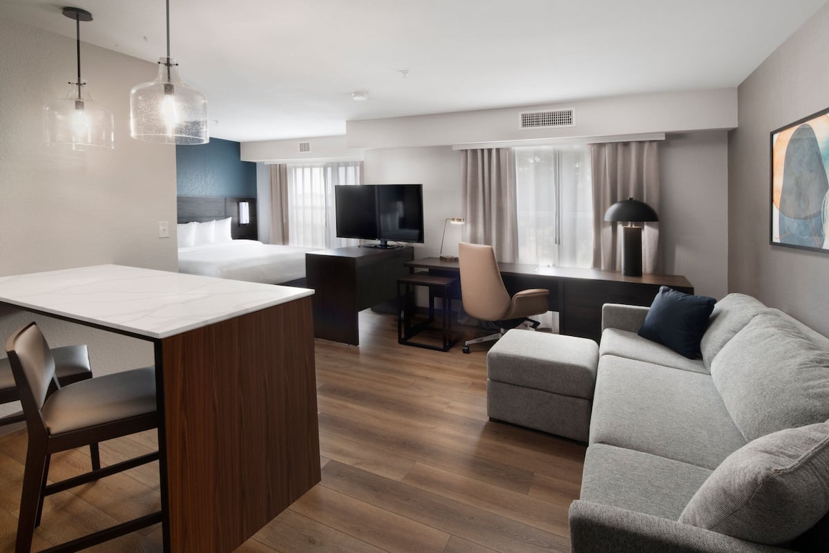 Una habitación de hotel moderna con piso de madera, un comedor con mesa y sillas blancas, un sofá gris, un televisor y un escritorio con silla.