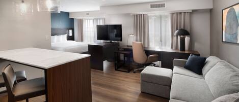 A modern hotel room with a wooden floor, a dining area with a white table and chairs, a gray sofa, a TV, and a desk with a chair.
