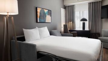 A hotel room with a bed, bedside table, lamp, chair, and a painting on the wall.