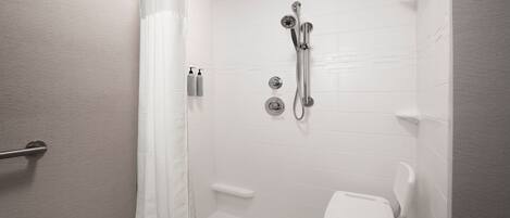 A walk-in shower with a white tiled wall, a showerhead, a handheld showerhead, a handheld shower attachment, a handheld shower seat, and a small shelf.