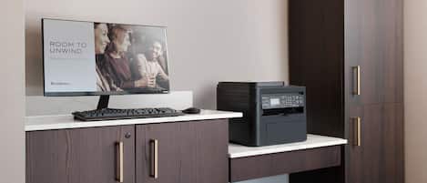 A modern office setup with a desktop computer, a printer, and a monitor displaying "ROOM TO UNWIND" on the screen.