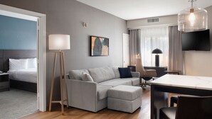 A modern hotel room with a grey sofa, a wooden floor, a flat-screen TV, and a dining table.