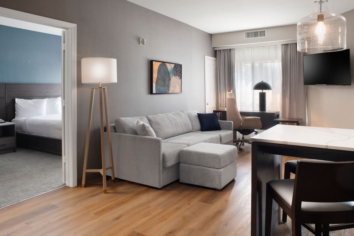 Una habitación de hotel moderna con un sofá gris, piso de madera, televisor de pantalla plana y una mesa de comedor.