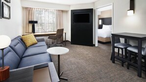 Quarto de hotel com sofá, mesa redonda pequena, escrivaninha, televisão na parede e cama com cabeceira.