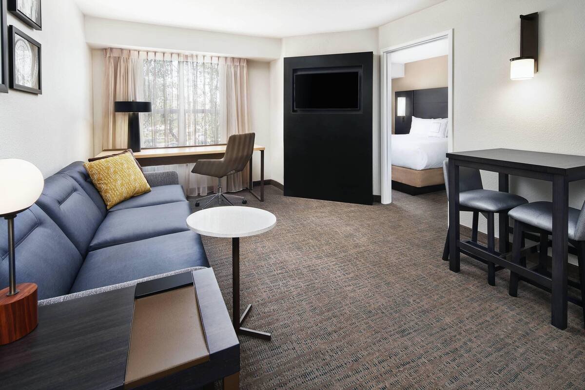 A hotel room with a sofa, a small round table, a desk, a TV mounted on the wall, and a bed with a headboard.