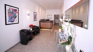 A reception area with a reception desk, a black leather sofa, a glass coffee table with magazines, and a potted orchid. - Hotel Savoy Bonn (Bonn)