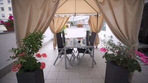 A covered outdoor seating area with a table and chairs, surrounded by potted plants and curtains. - Hotel Savoy Bonn (Bonn)