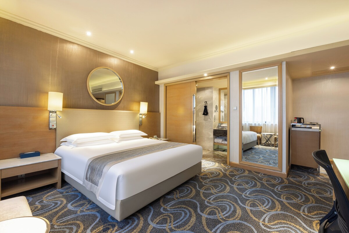 Une chambre d’hôtel avec un grand lit, des lampes de chevet, un miroir, un bureau et une salle de bain visible à travers une porte ouverte.