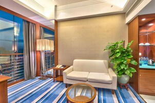 Ein Hotelzimmer mit einer Sitzecke, einem kleinen runden Tisch, einer Zimmerpflanze, einer Minibar und einem großen Fenster mit Vorhängen.