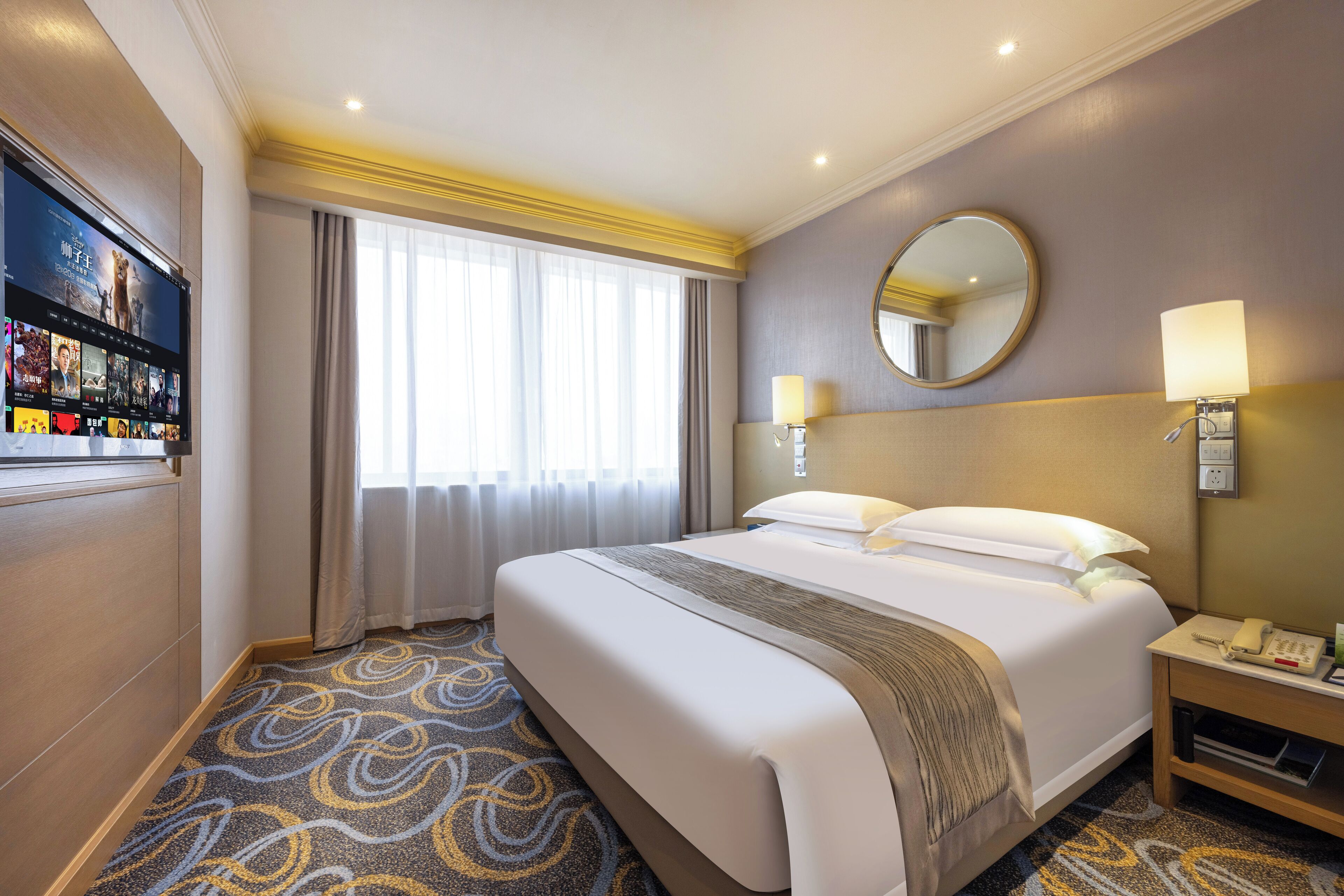 A hotel room with a large bed, a flat-screen TV mounted on the wall, a bedside table with a lamp, and a carpeted floor.