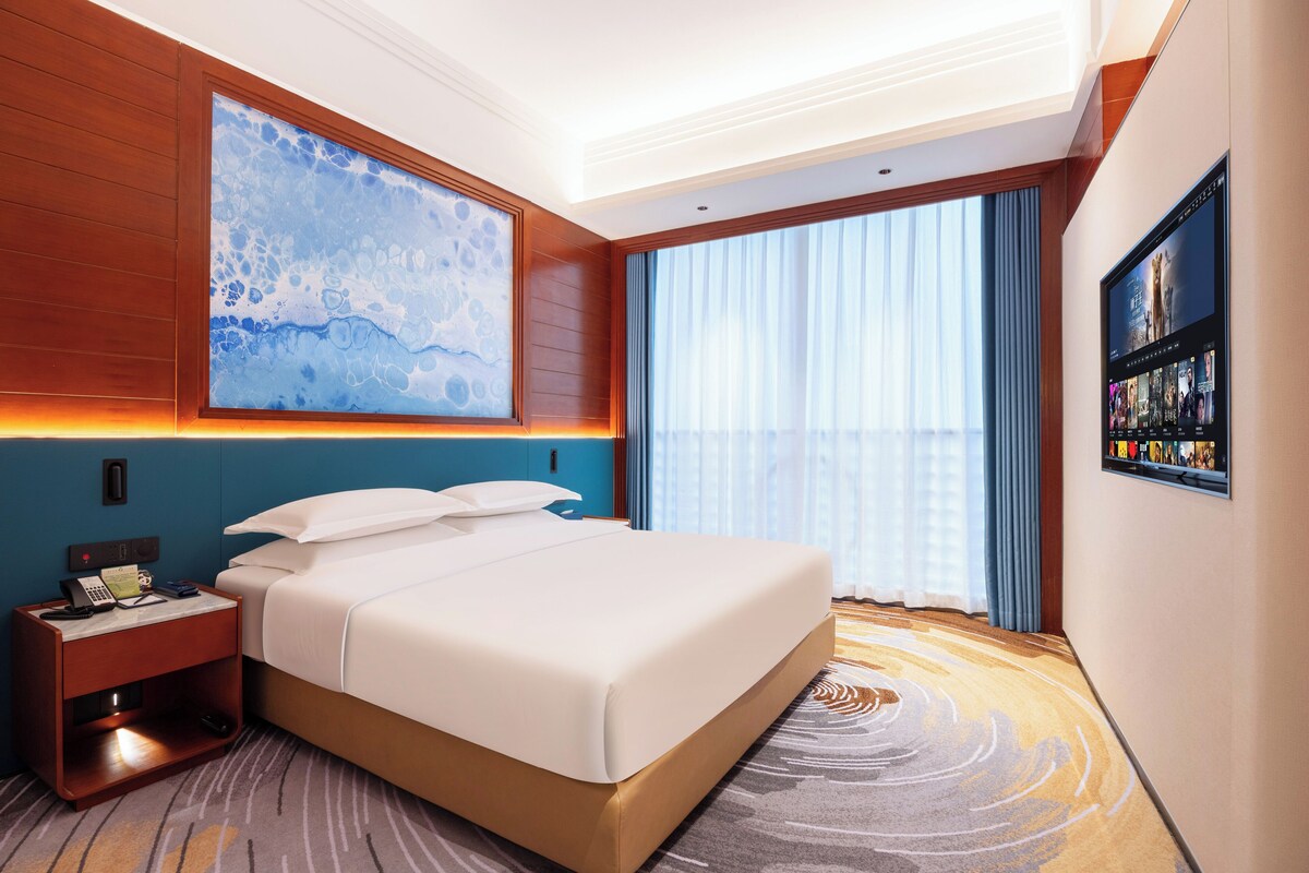 Une chambre d’hôtel moderne avec un grand lit, une table de chevet avec un téléphone et une lampe, une grande fenêtre avec des rideaux et un téléviseur fixé au mur.