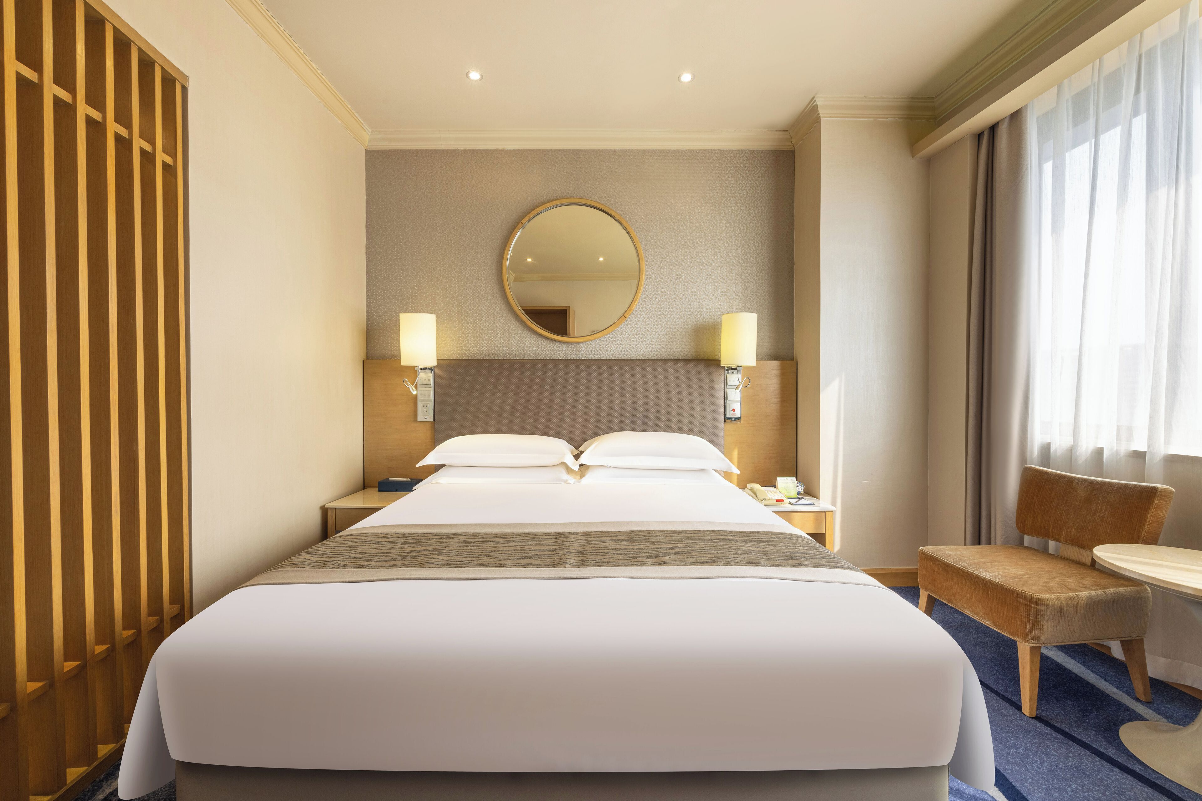 Une chambre d’hôtel avec un grand lit, deux tables de chevet avec des lampes, un grand miroir rond au mur, une armoire en bois et une petite table ronde avec une chaise.