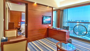 Habitación de hotel con pared de paneles de madera, televisor, escritorio con silla, una mesita pequeña y un ventanal grande con vistas a la ciudad.
