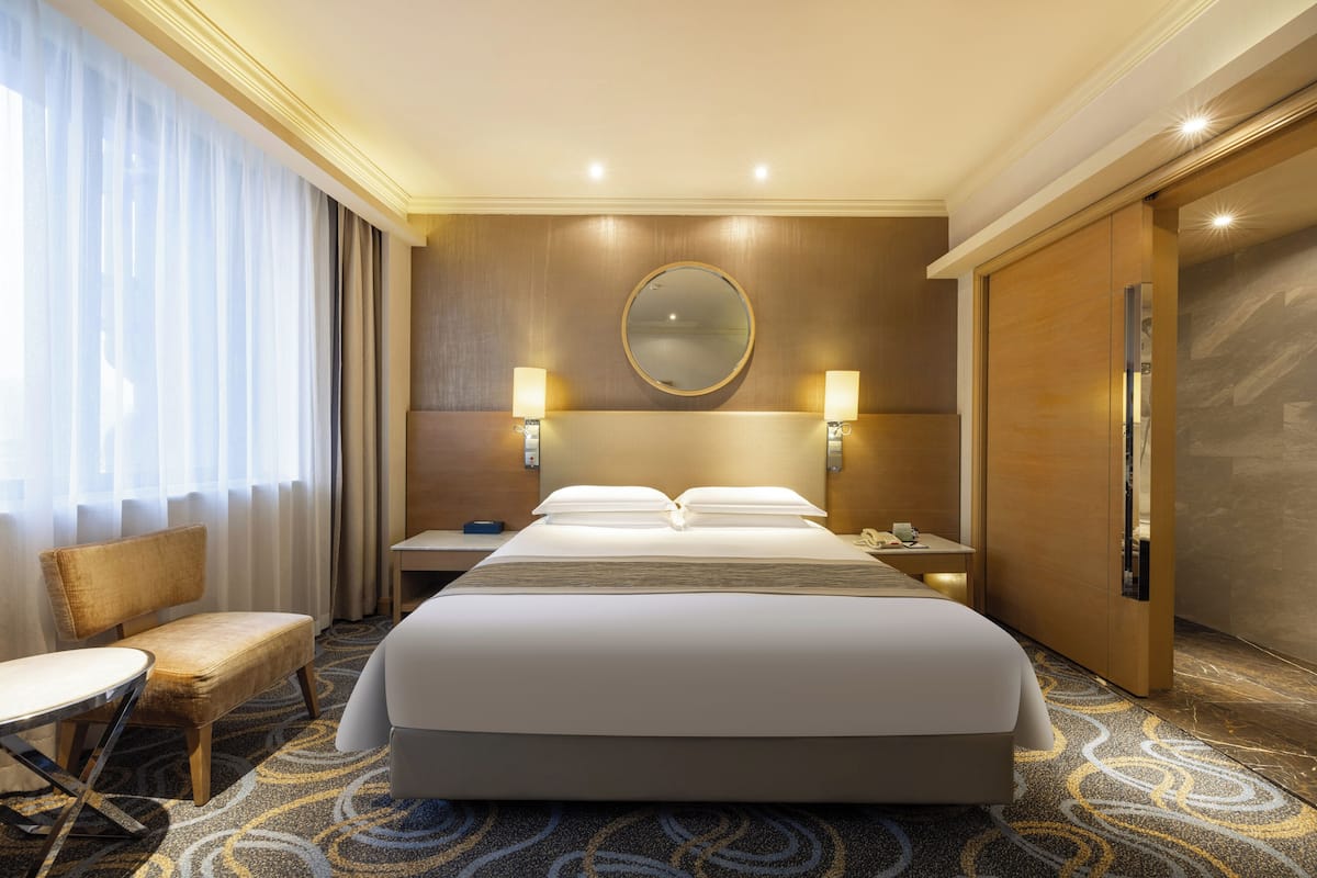 Quarto de hotel com uma cama grande, duas mesas de cabeceira, uma cadeira, uma escrivaninha, um espelho e uma janela com cortinas.