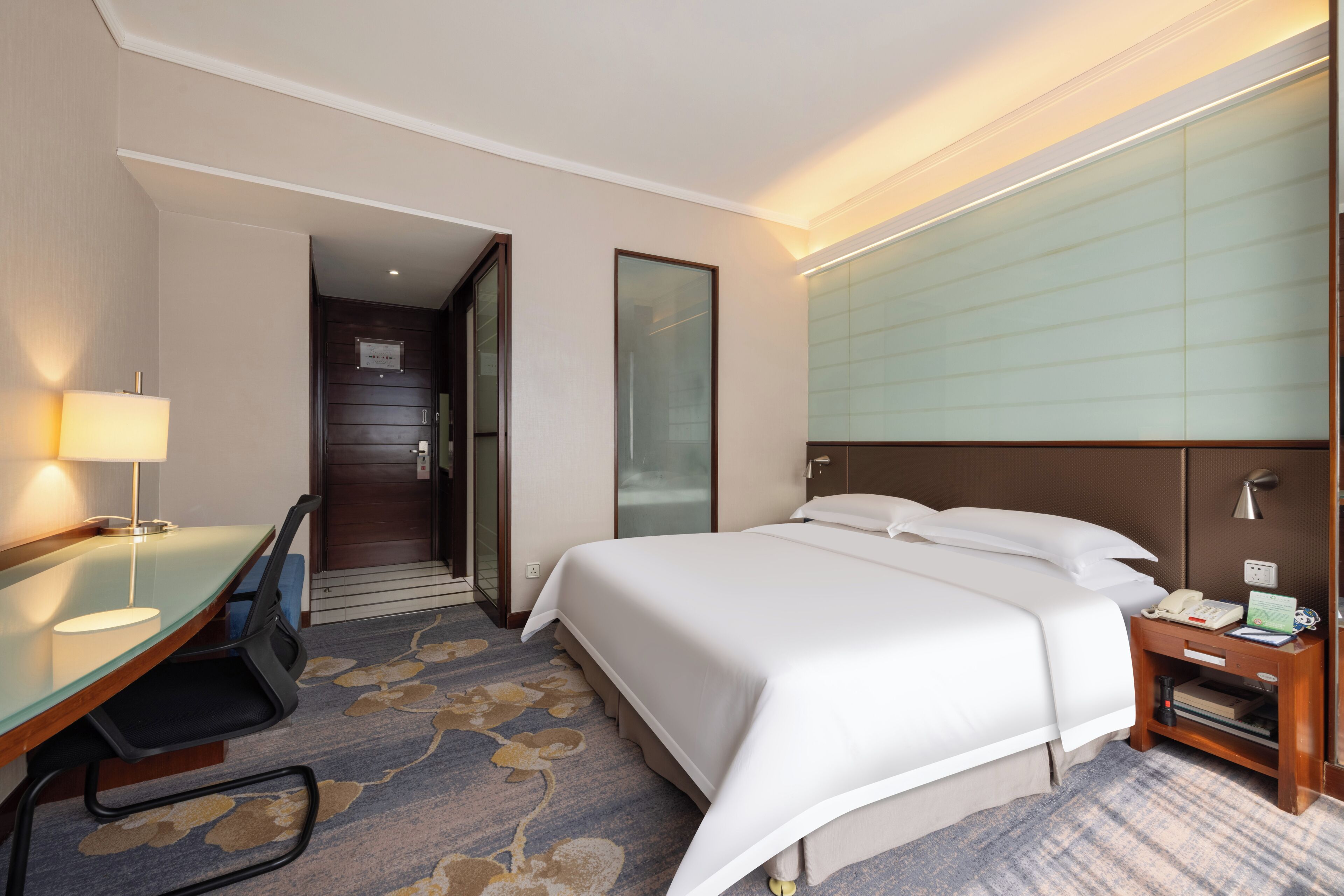 A hotel room with a bed, desk, chair, lamp, bedside table, and a door leading to another room.