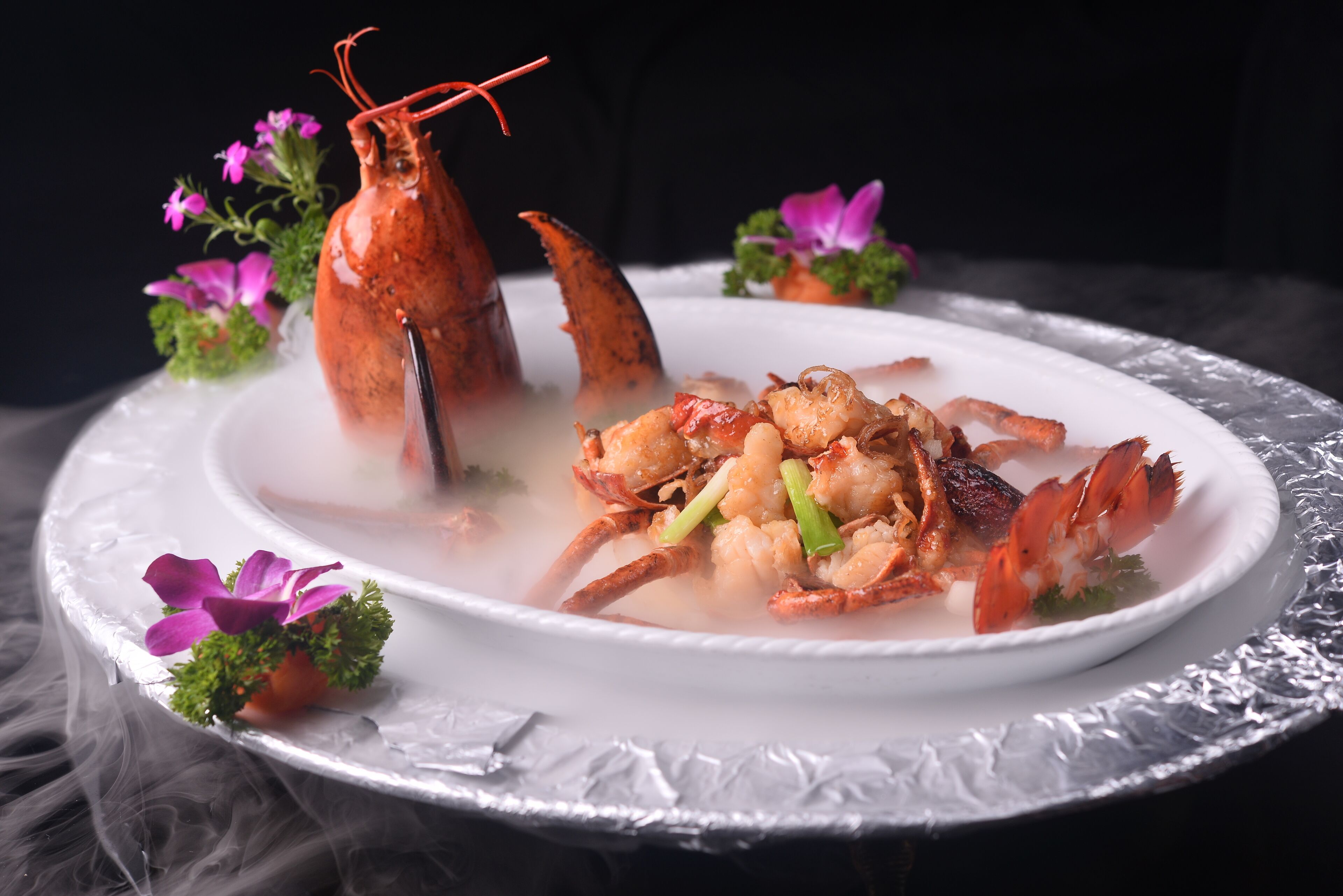 A seafood dish with lobster and shrimp on a white plate, surrounded by purple flowers and green garnish.