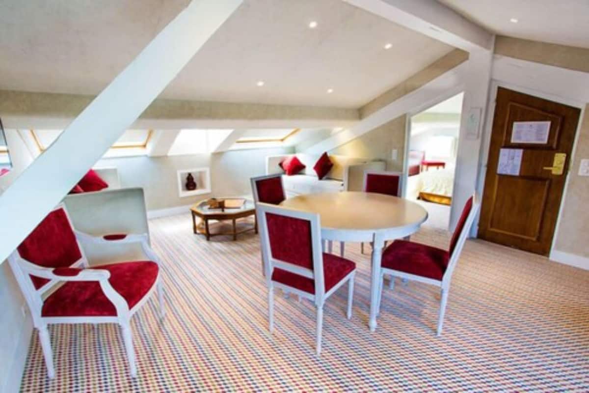 A room with a high ceiling, a round table, and red chairs. There is a bed with red pillows and a small table with a chair.