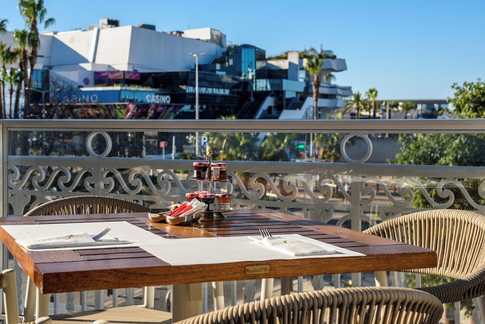 Une table en bois, sur laquelle sont disposés des condiments, un menu et une bouteille, se trouve sur un balcon offrant une vue sur un casino et des palmiers.
