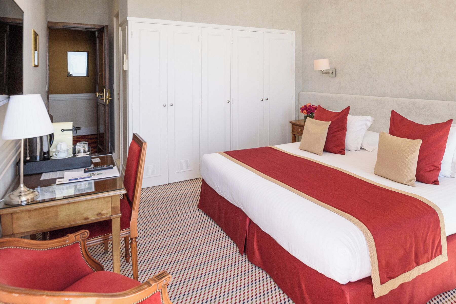 A hotel room with a bed, a desk, a chair, a wardrobe, and a nightstand with a lamp.