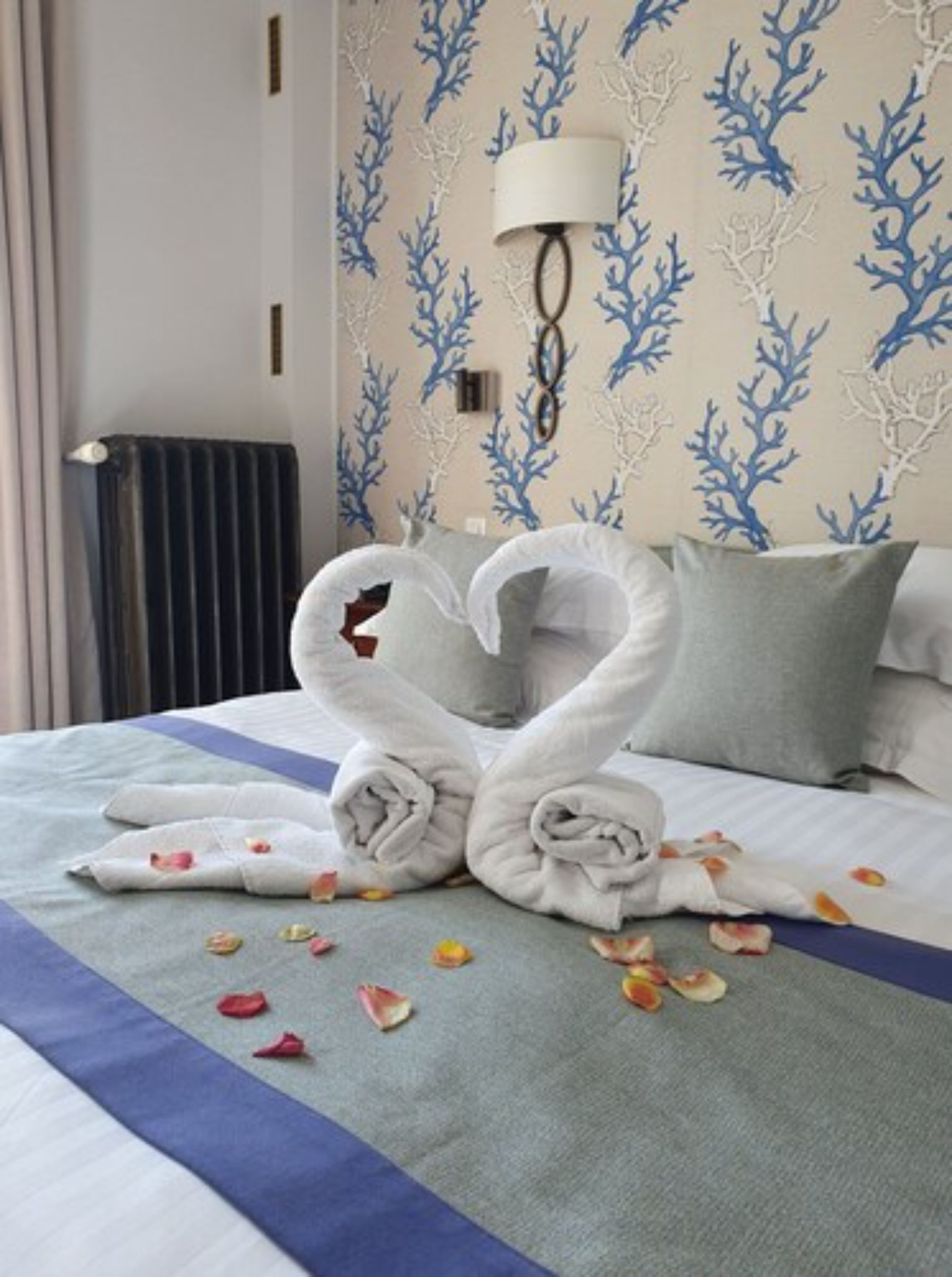 A neatly made bed with towels arranged in a heart shape, a lamp, and a wall with a blue and white pattern.