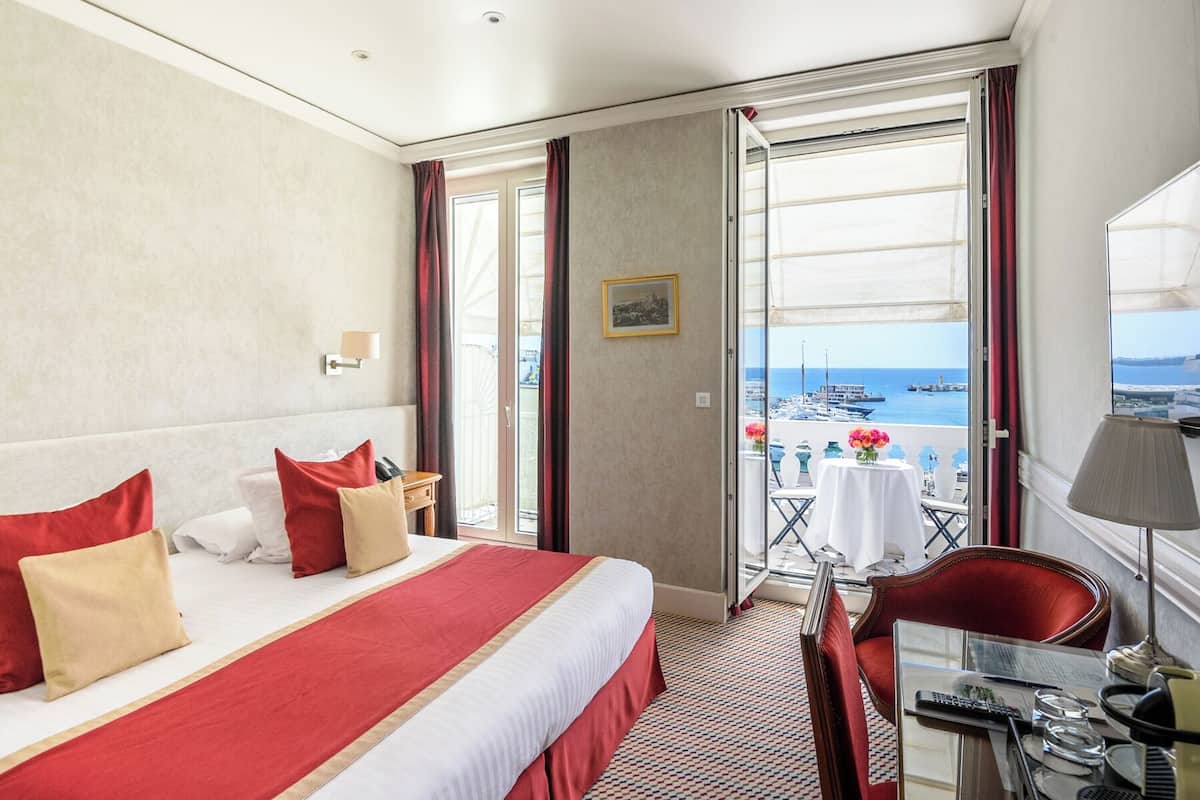 A hotel room with a bed, a desk, a chair, a table with a vase, and a view of the sea.