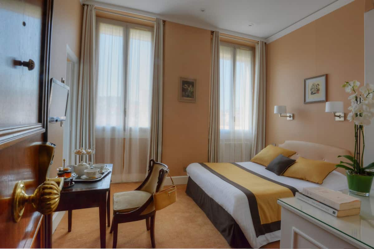 A hotel room with a bed, a desk with a chair, a small table with a tray of items, and a potted plant.