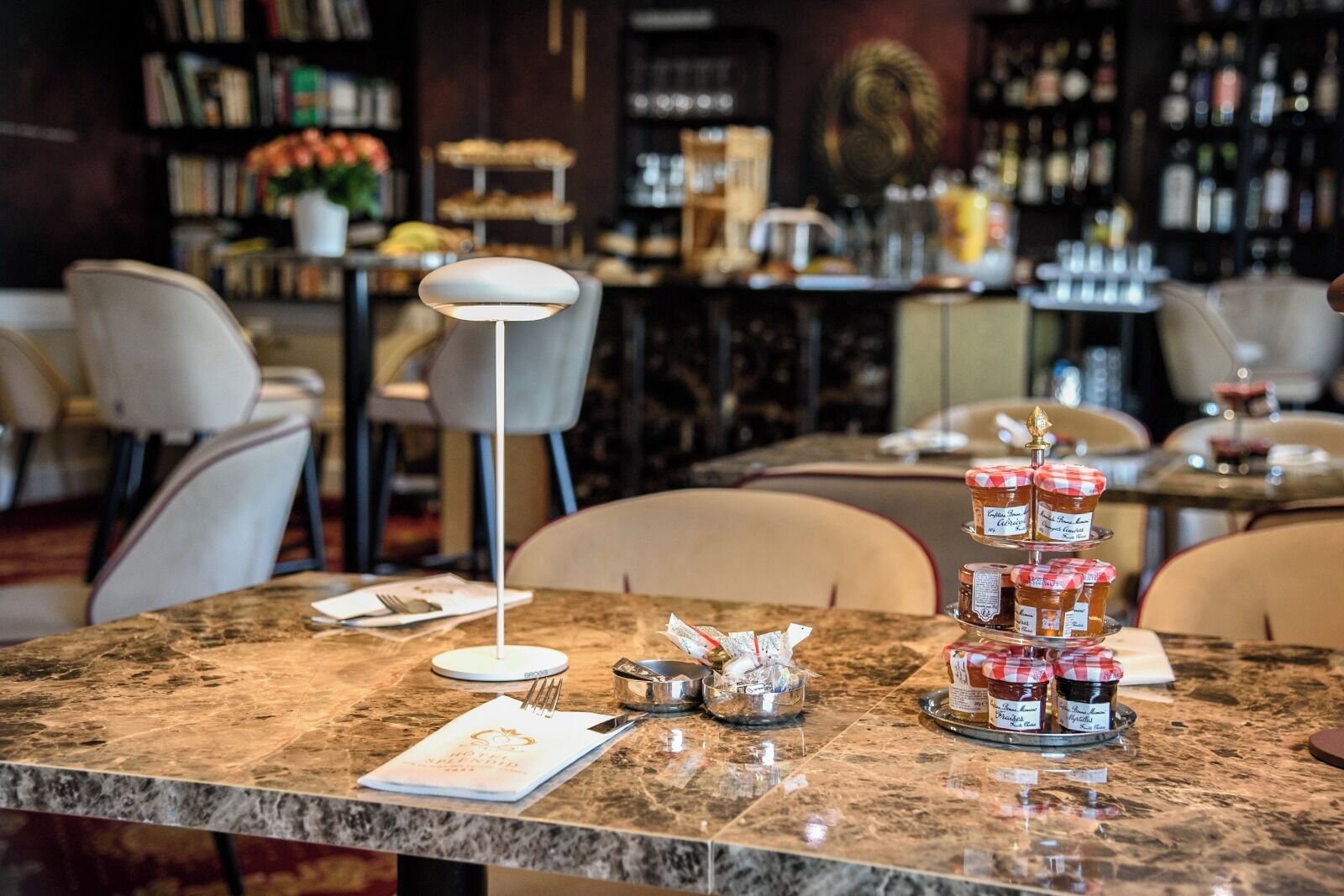 Interior de restaurante con mesas de mármol, una mesa de postres escalonada y una barra de bar al fondo.