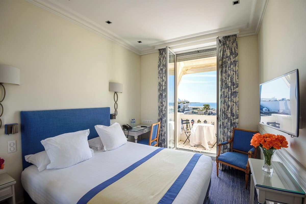 A hotel room with a large bed, a desk, a chair, a TV, and a balcony with a view.