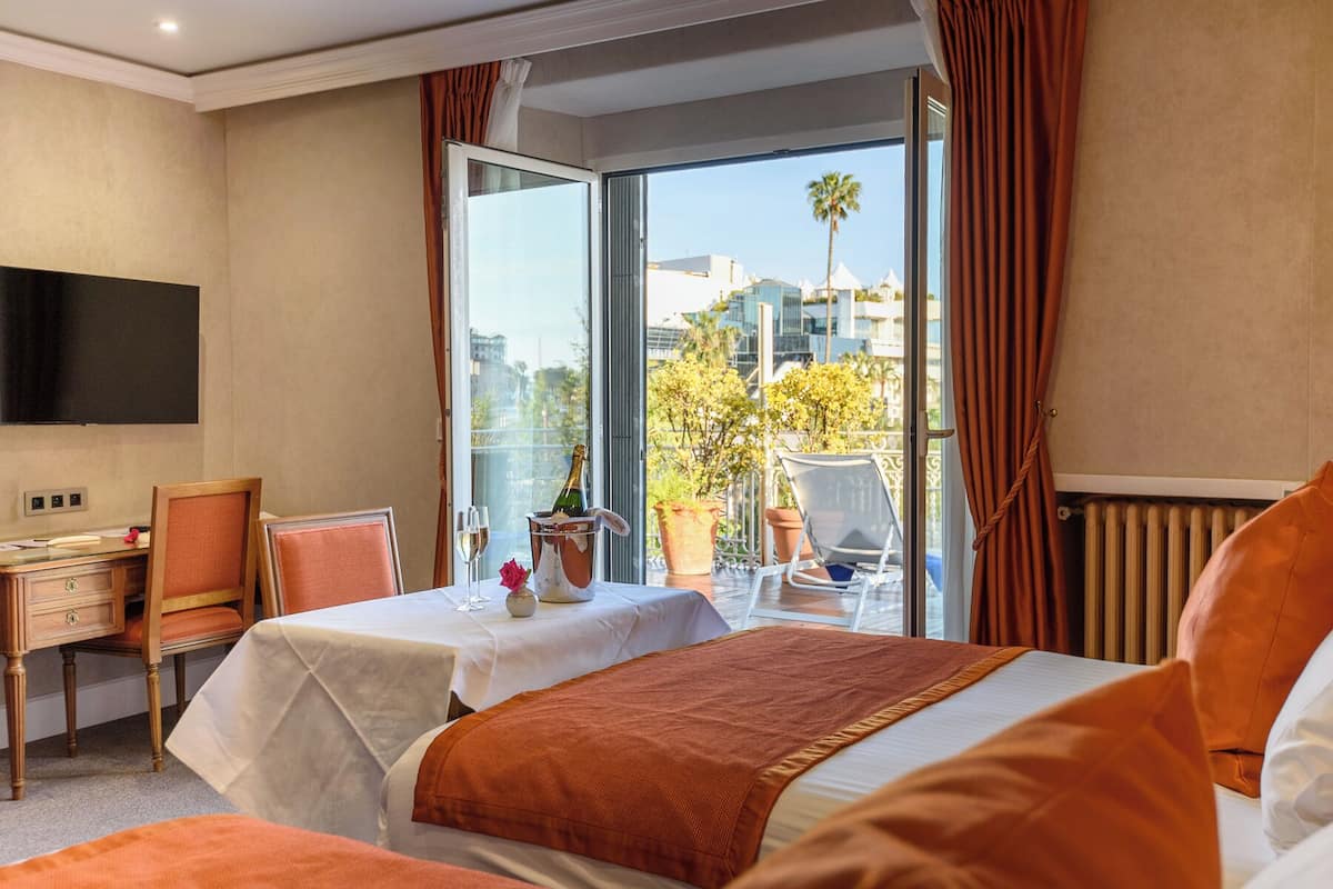 A hotel room with a bed, a desk, a chair, a TV, and a balcony with a view of buildings and trees.