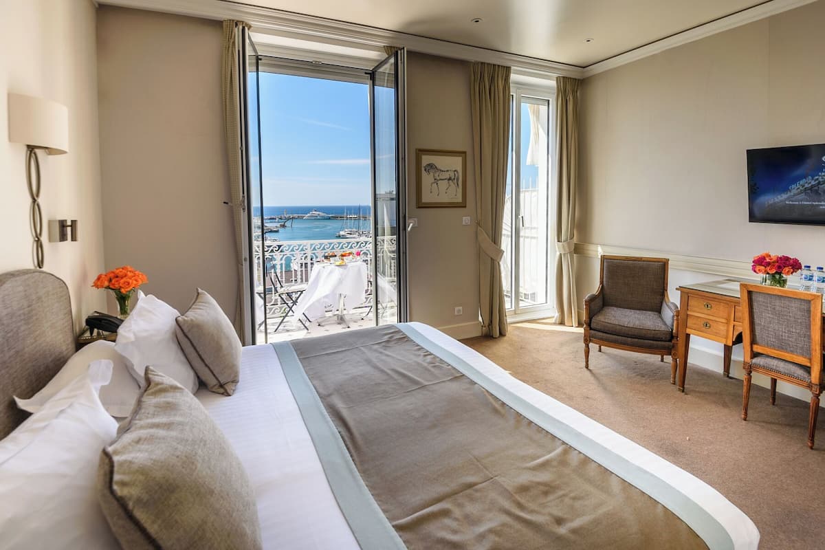 A hotel room with a bed, a chair, a desk, a lamp, a picture on the wall, and a view of the sea and boats.